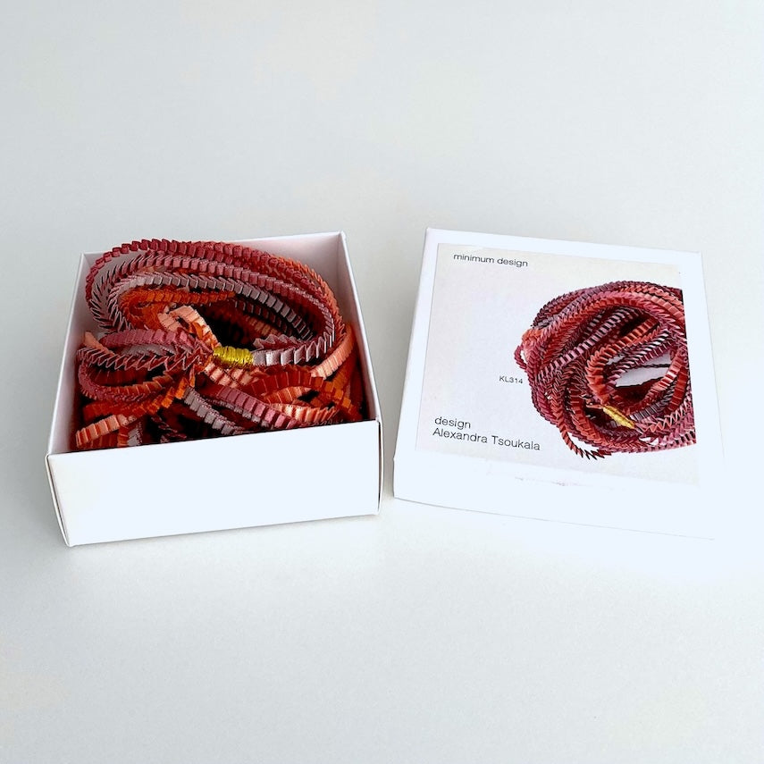 Long collier composé de fines bandes de tissus colorés et plissés est présenté dans une boîte en carton blanc illustrée de la marque de la créatrice grecque, Alexandra Tsoukala.