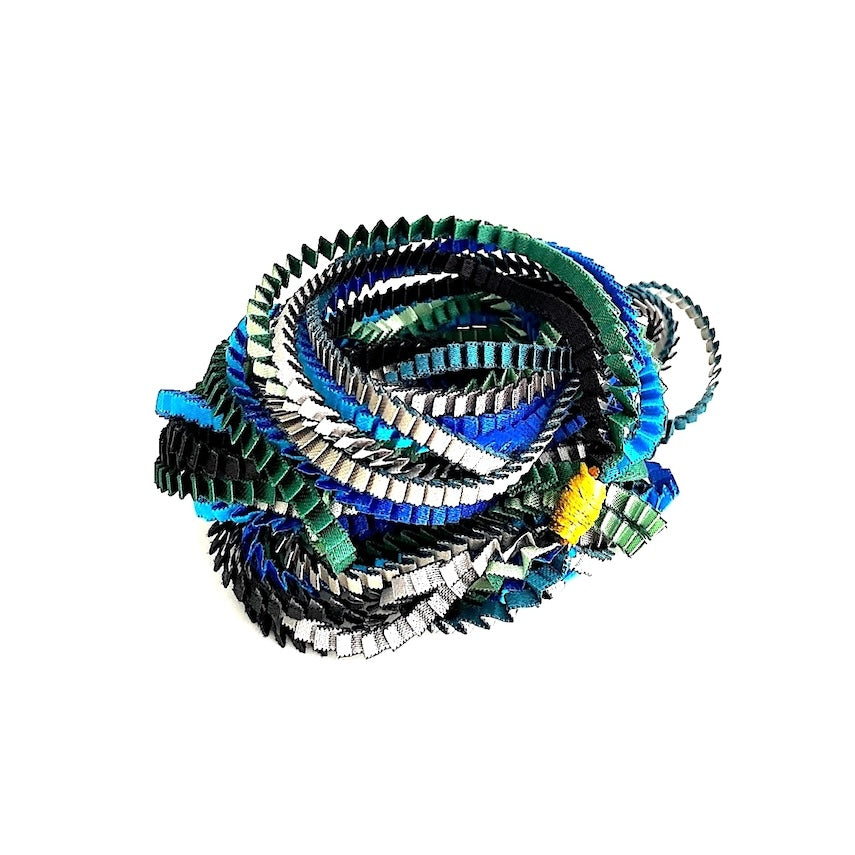 Long collier façonné de huit bandes de tissus plissés et satinés de couleurs : bleu, vert foncé, turquoise, argenté, noir. Chaque bandes est réliées par un lien de coton jaune.