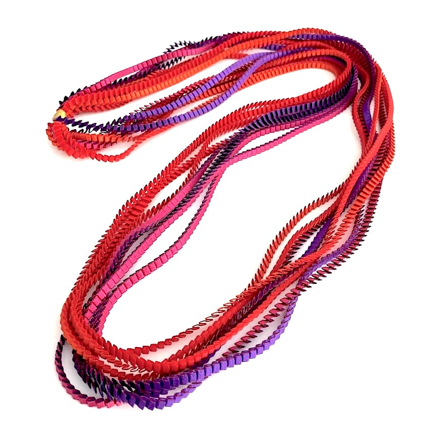 Long sautoir coloré composé de huit bandes de satin plissé rouge, orange, violet, fuchsia reliées par un lien de coton jaune d'or. Ce collier se porte aussi en double.