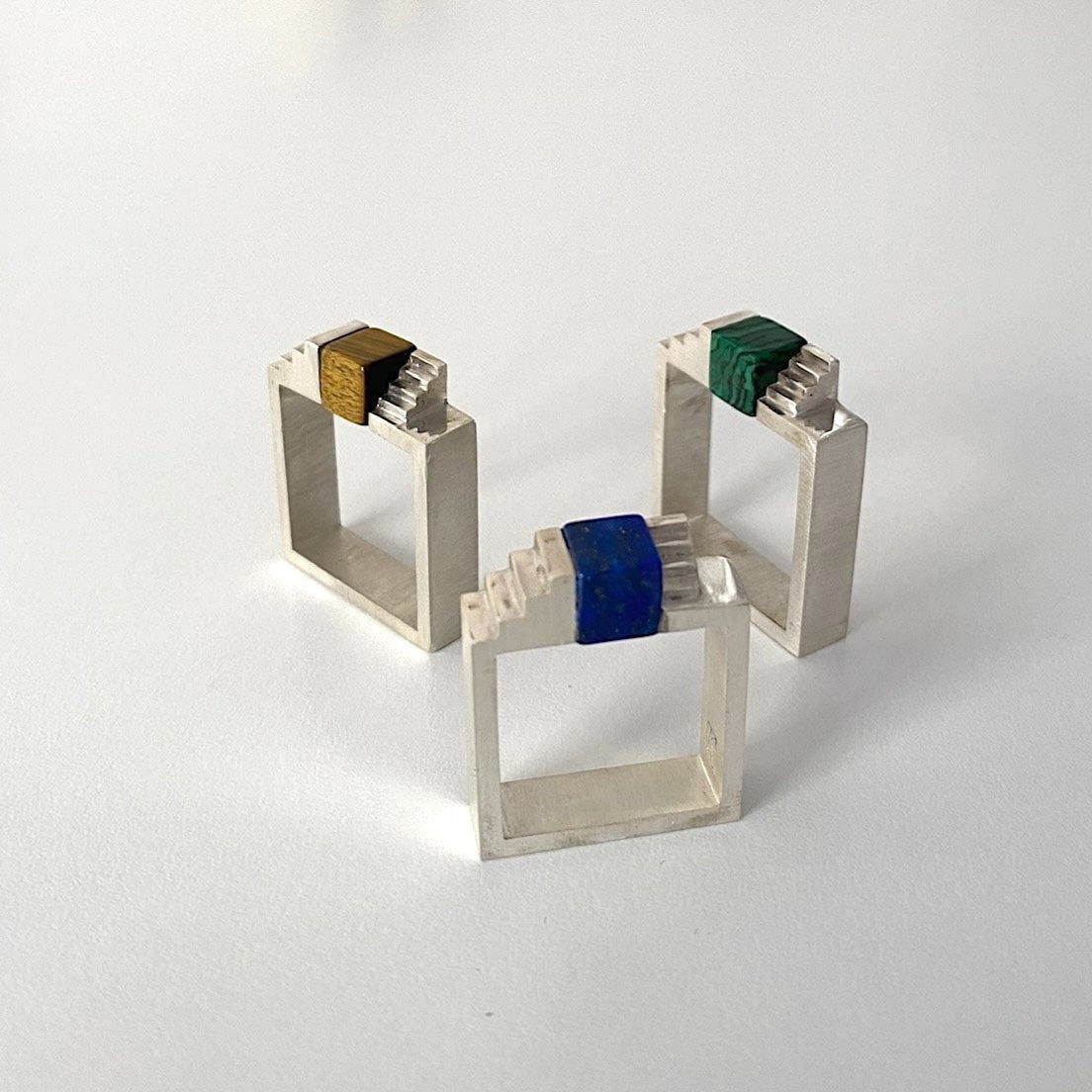 Trois bagues géométriques en argent brossé. Chaque bague est sertie d'un cube de gemmes différentes : un lapis-lazuli, un oeil de tigre et d'une malachite.