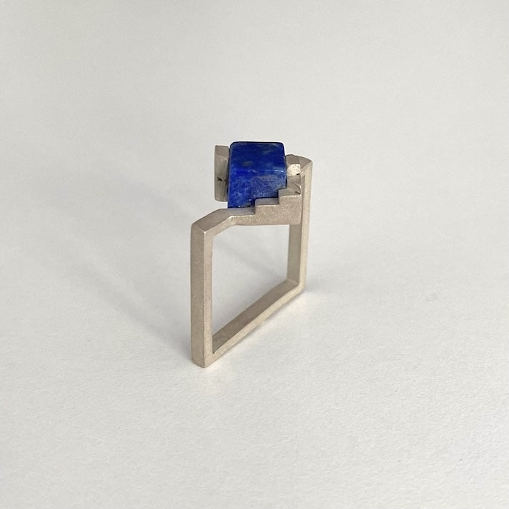 Bague géométrique composée d'un anneau fin et carré en argent brossé sertie un cube de lapis-lazuli imbriqué entre deux éléments en formes d'escalier.