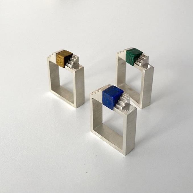 Trois bagues géométriques en argent brossé. Chaque bague est sertie d'un cube de gemmes différentes : un lapis-lazuli, un oeil de tigre et d'une malachite.