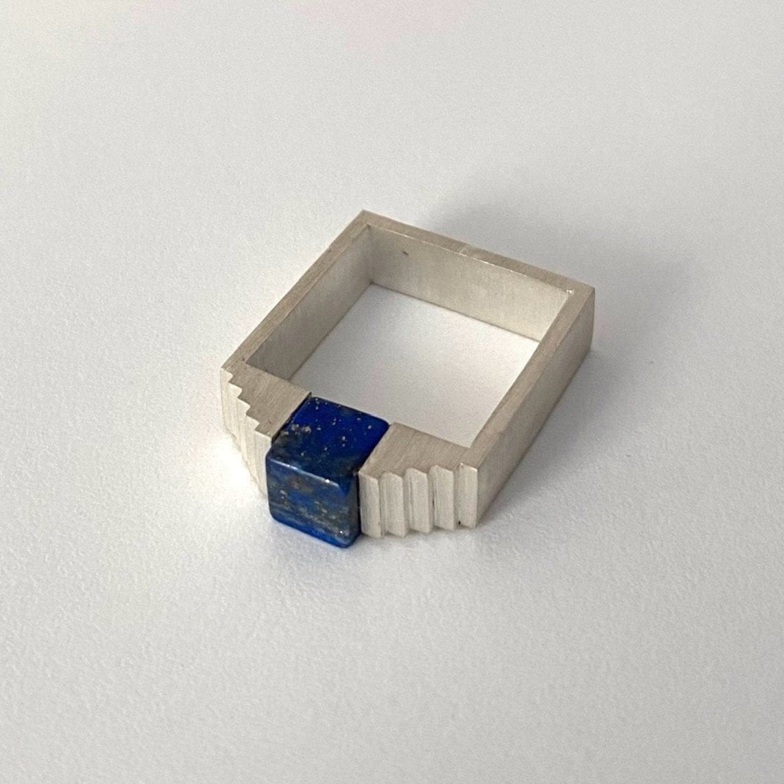 Bague géométrique en argent brossé avec un cube de lapis-lazuli imbriqué entre deux éléments en formes d'escalier.