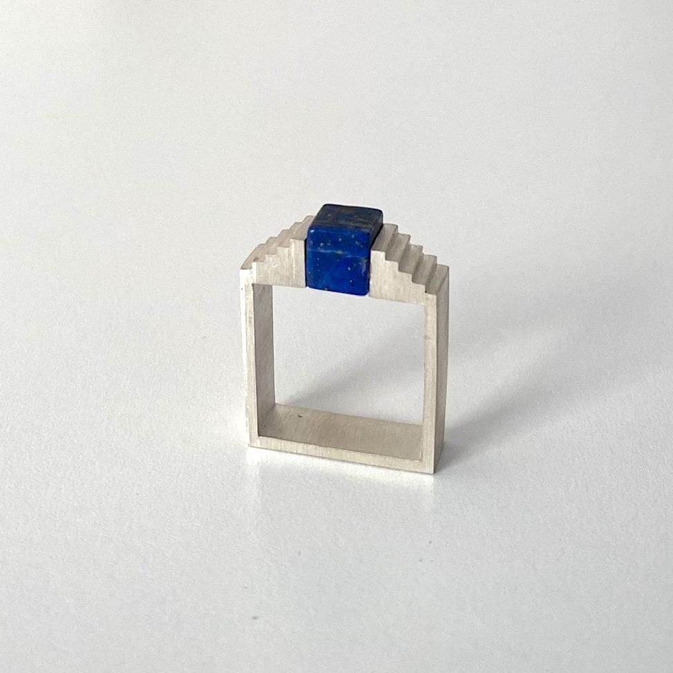 Bague architecturale en argent brossé avec un cube de lapis-lazuli imbriqué entre deux éléments en formes d'escalier.