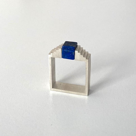 Bague architecturale en argent brossé avec un cube de lapis-lazuli imbriqué entre deux éléments en formes d'escalier.