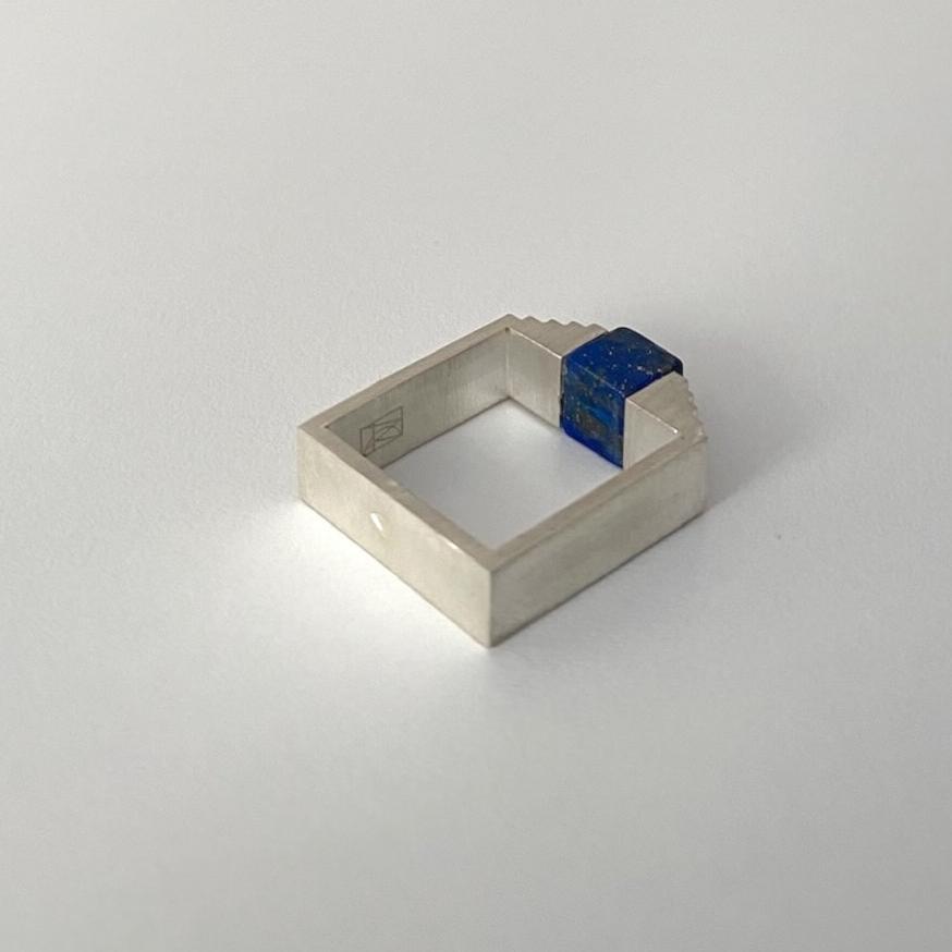 Bague architecturale en argent brossé. Sur le dessus de l'anneau carré, un lapis-lazuli taillé en cube est imbriqué entre deux éléments en formes d'escalier.