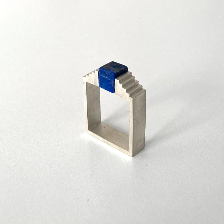 Bague carrée en argent brossé. Sur le dessus de l'anneau carré, un lapis-lazuli taillé en cube est imbriqué entre deux éléments en forme d'escalier.