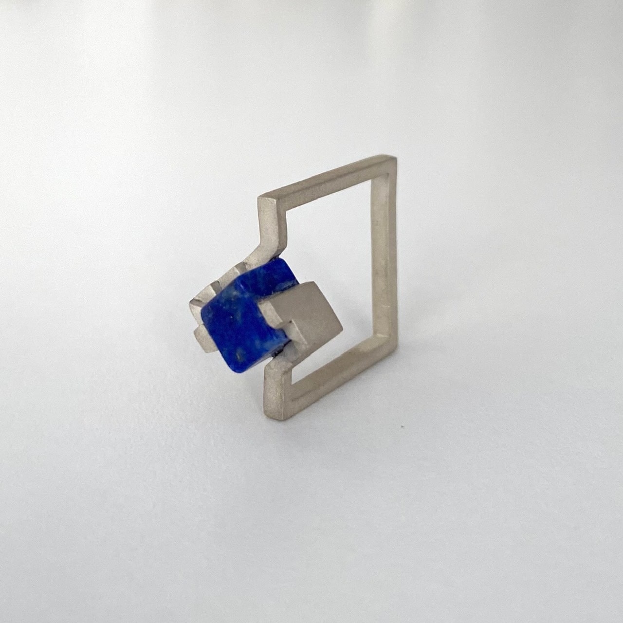 Bague solitaire, anneau fin et carré en argent brossé avec un cube de lapis-lazuli imbriqué entre deux éléments en formes d'escalier.