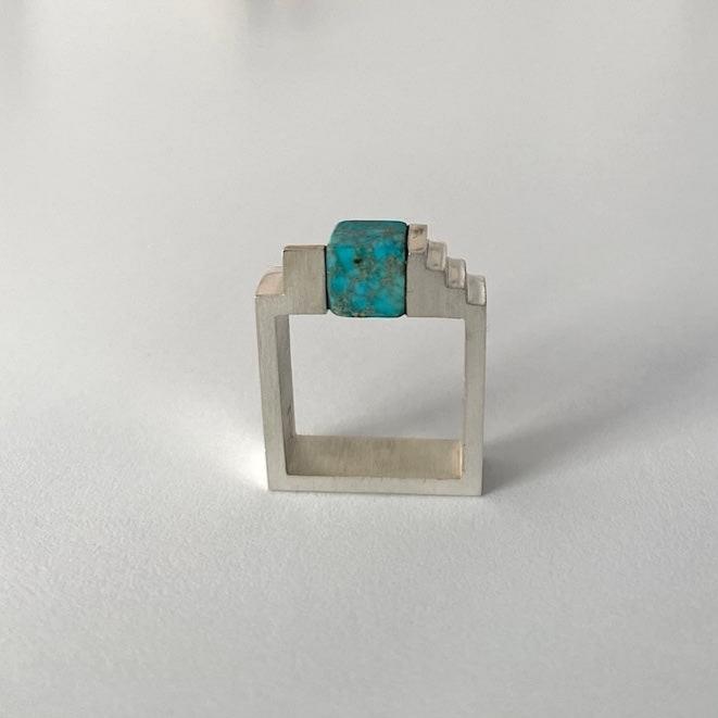 Bague géométrique en argent brossé, au centre une turquoise taillée en cube est enchâssée entre deux éléments inversés en forme d'escalier.