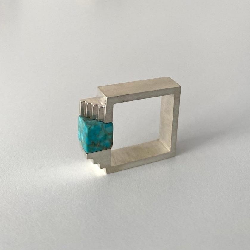 Bague carrée en argent brossé, au centre une turquoise taillée en cube est enchâssée entre deux éléments inversés en forme d'escalier.