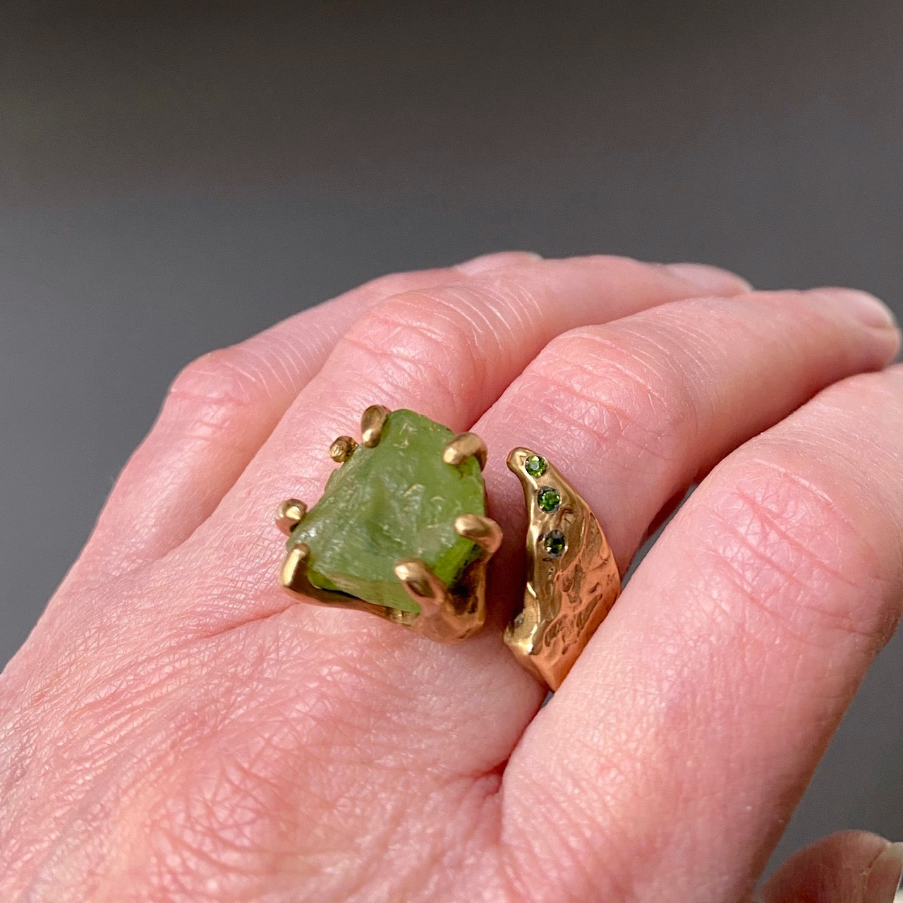 Bague ouverte sculpturale en bronze avec péridot brut maintenu par un serti griffes, sur le côté 3 Diopsides verts. Détail main.