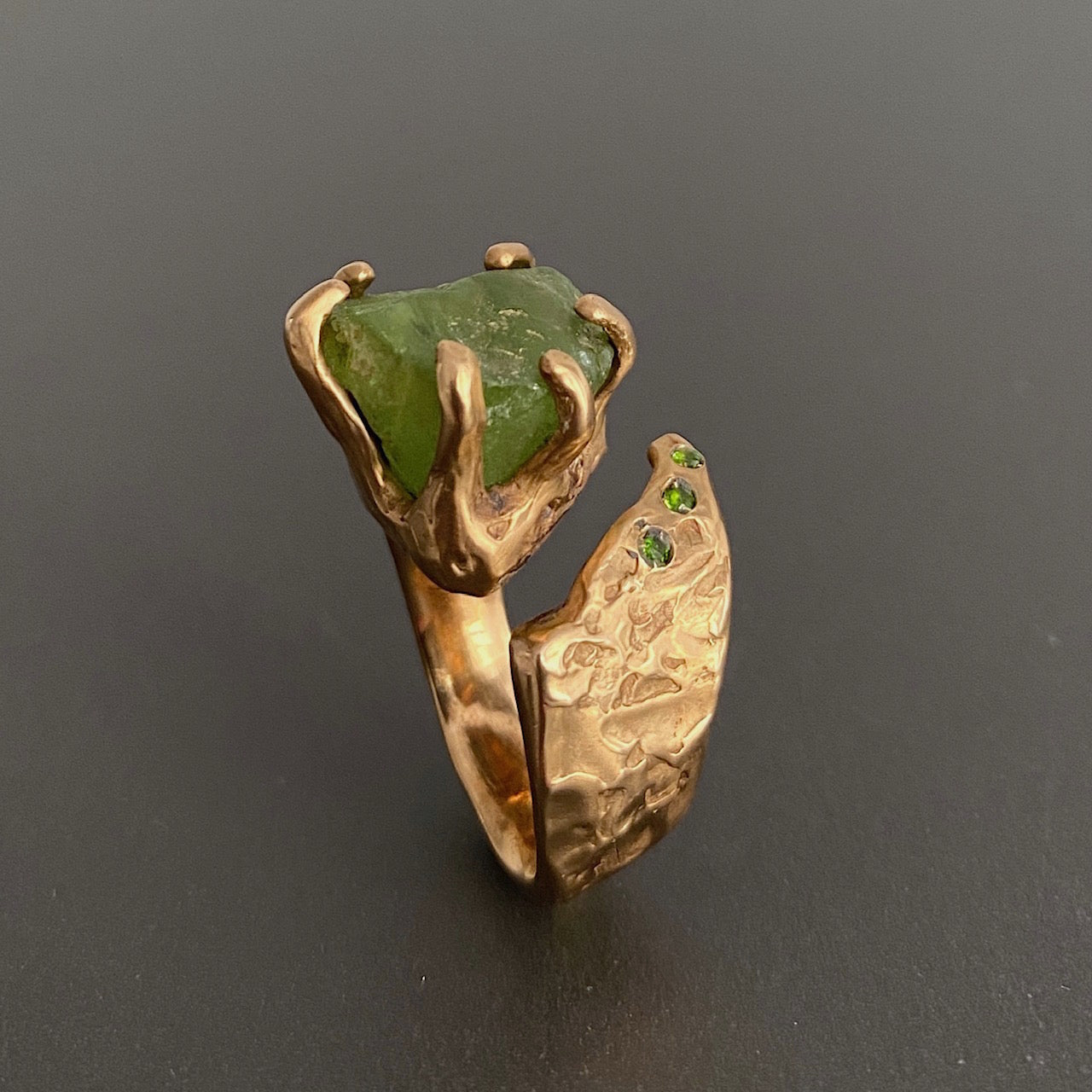 Bague ouverte sculpturale en bronze avec péridot brut maintenu par un serti griffes, sur le côté 3 Diopsides verts. Bague debout sur fond gris.