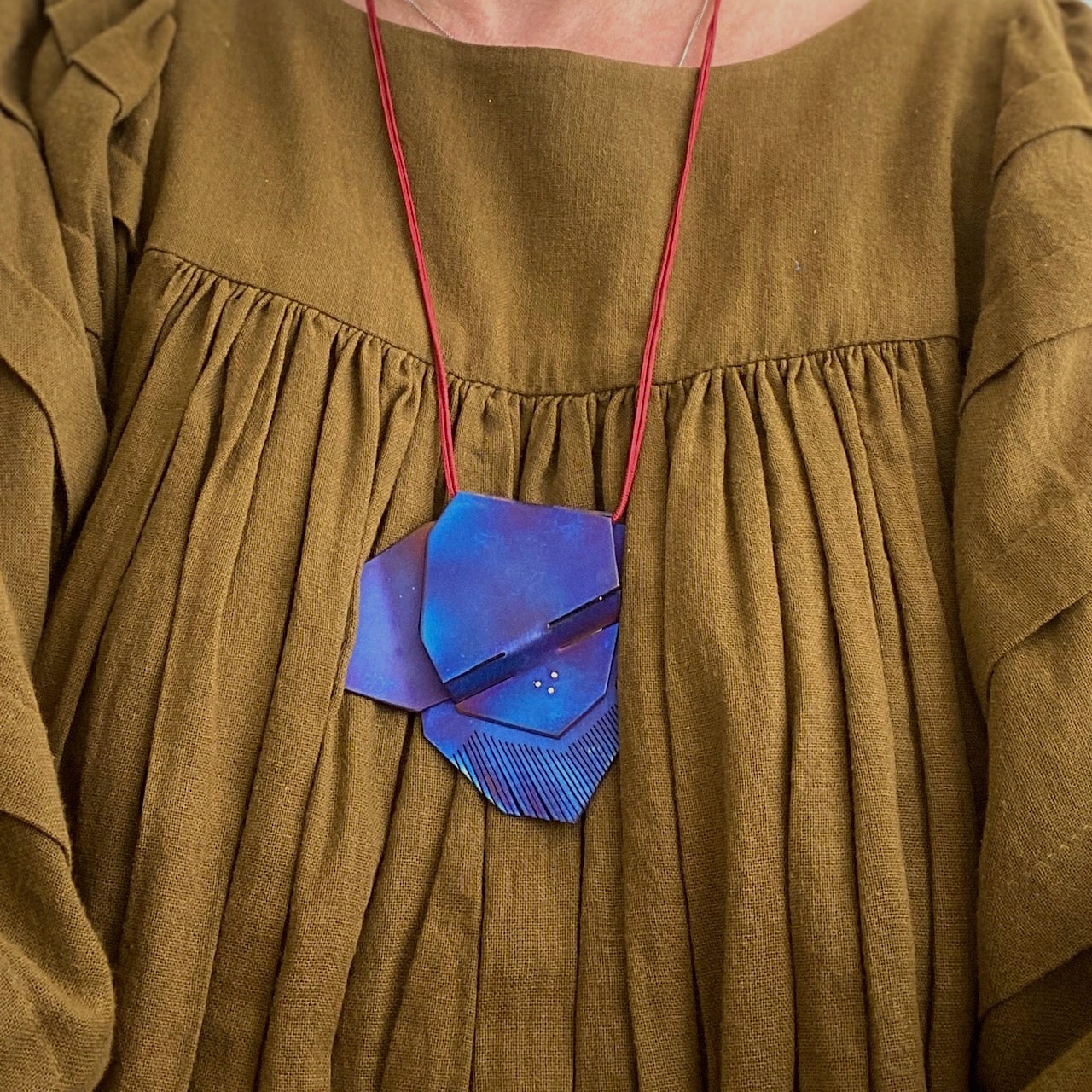 Pendentif sculptural en plaques de titane bleuï, pliées et sciées. Bijou porté sur chemise kaki.i.