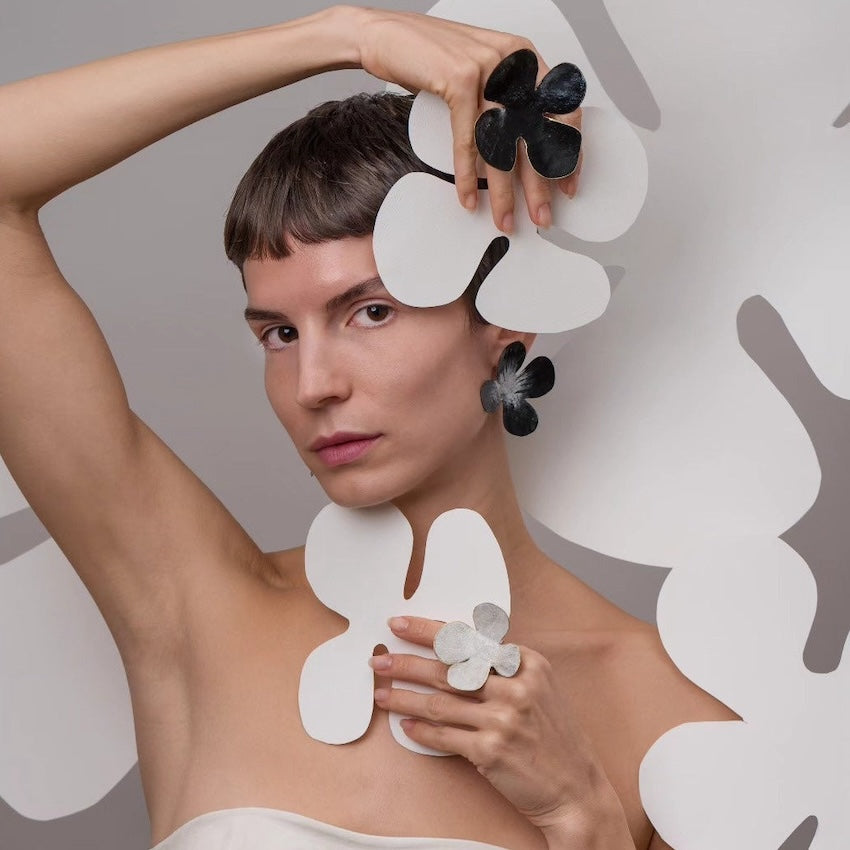 Portrait de femme dans un décor florale, elle porte une boucle en forme de fleur noire et une bague fleur à chaque main.