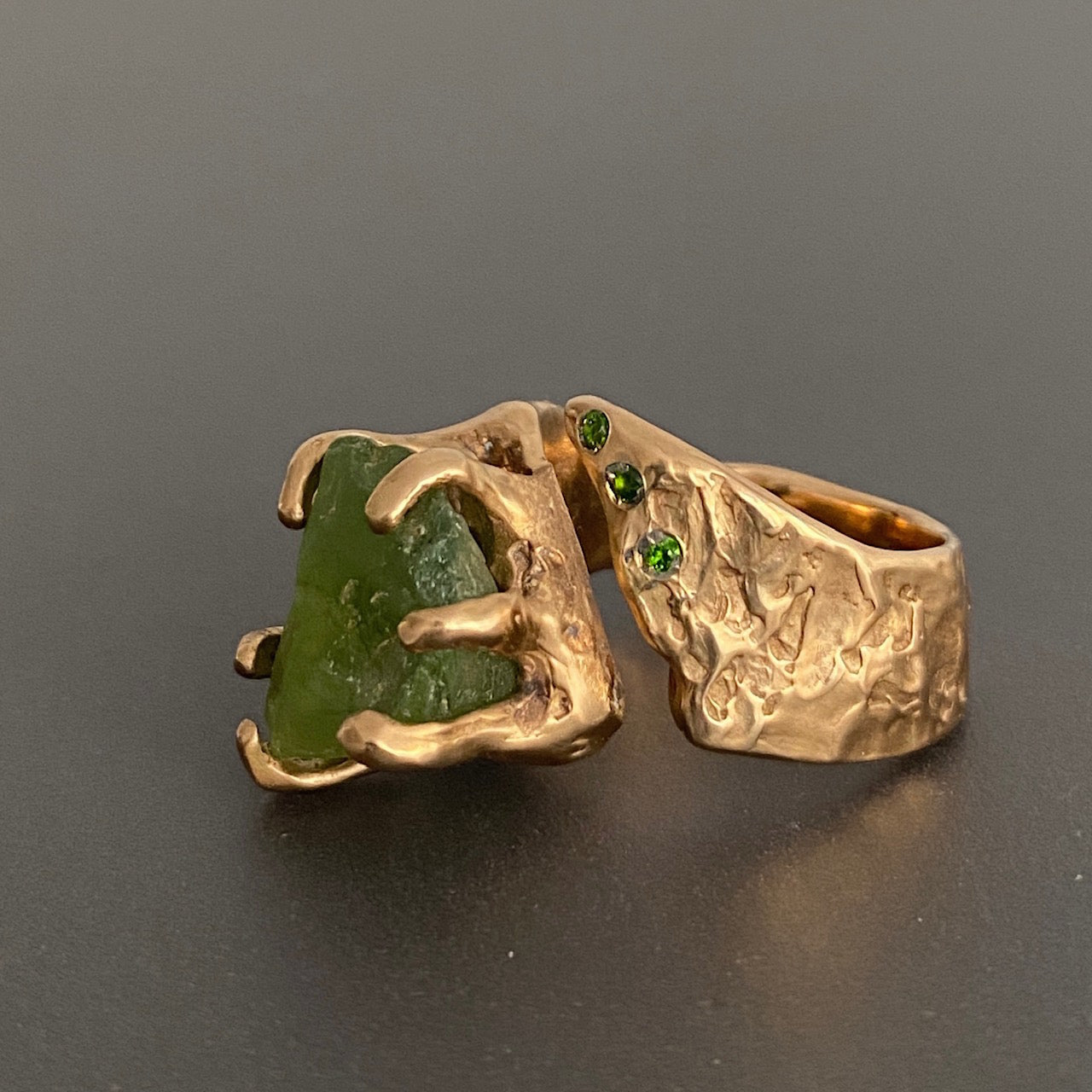 Bague ouverte sculpturale en bronze avec péridot brut maintenu par un serti griffes, sur le côté 3 Diopsides verts. Bague posée sur fond gris.