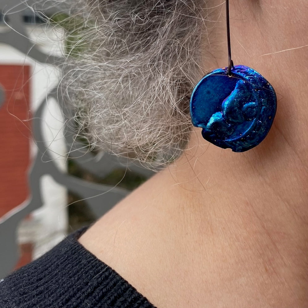 Boucle pendante en titane bleu et texturé en forme de disque irrégulier présenté sur une tige droite en titane noir. Détail profil et cou.